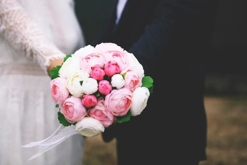 Novios tomados del brazo; ella sostiene un ramo de peonías rosas y blancas.